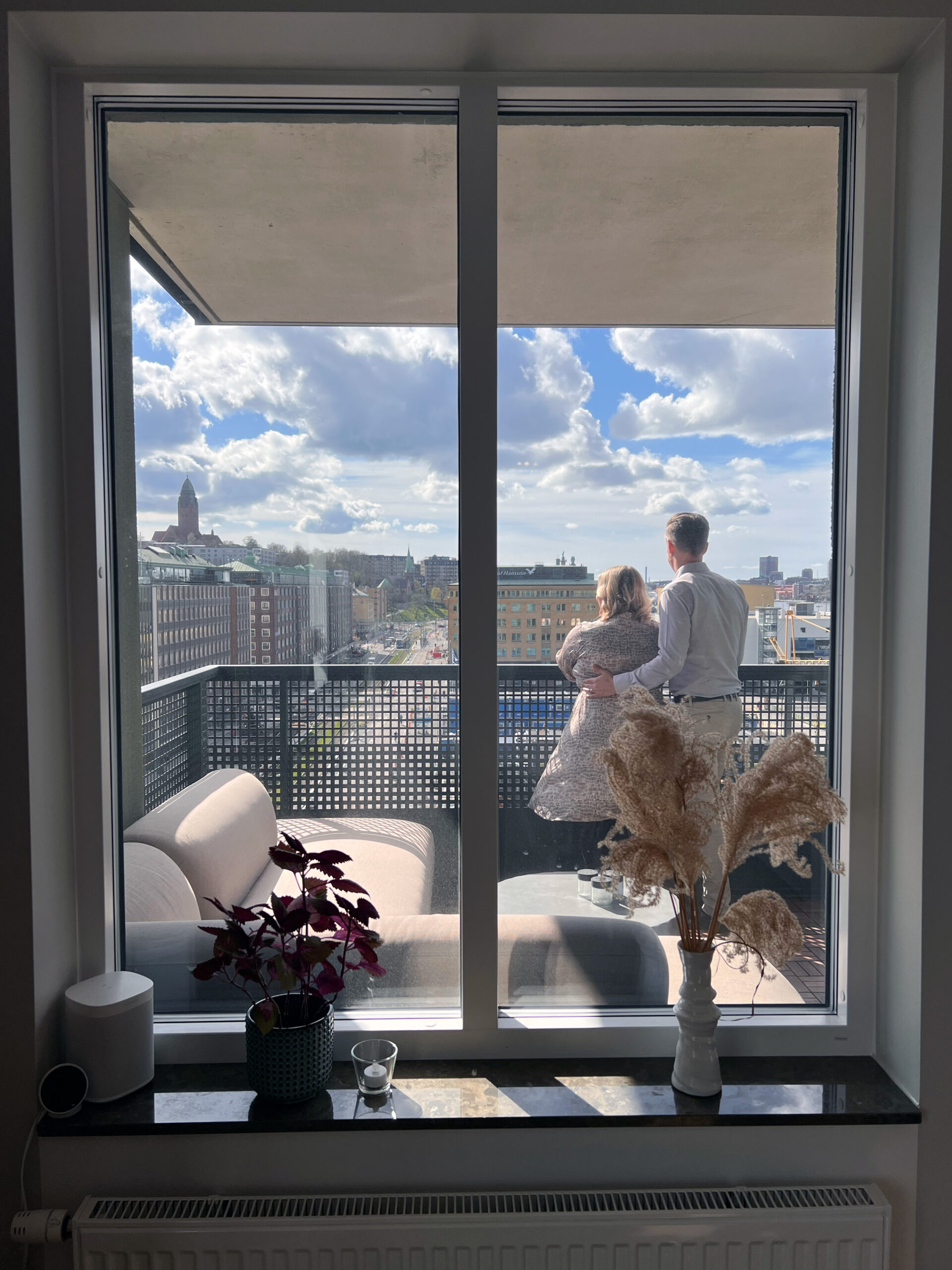 Två personer står på en balkong. De är fotade genom ett stängt fönster.